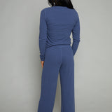 A woman viewed from behind wearing a matching slate blue long-sleeve top and wide-leg pants set, standing against a plain light gray studio background.