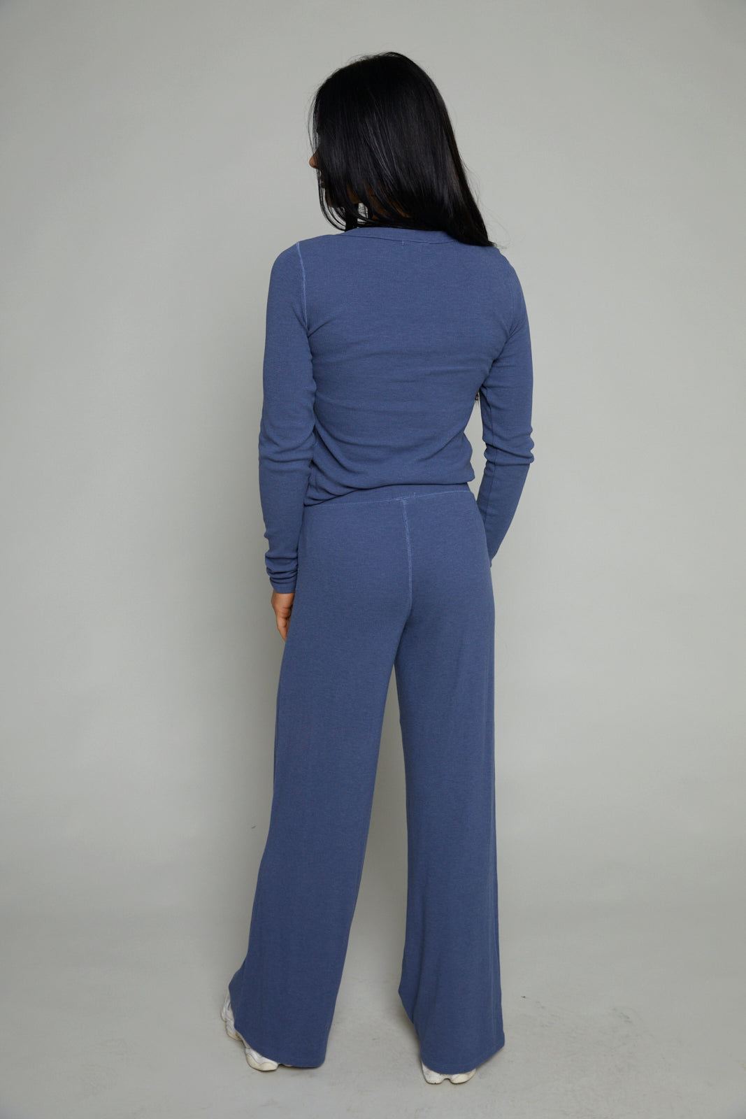 A woman viewed from behind wearing a matching slate blue long-sleeve top and wide-leg pants set, standing against a plain light gray studio background.